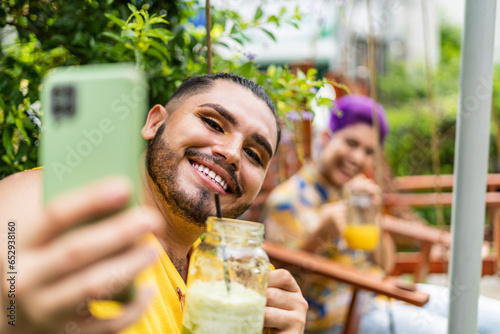 portrait of a homosexual man with makeup taking a very happy picture with his cell phone and having a drink and behind him his short-haired friend with purple hair smiling  photo