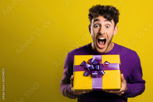 Handsome surprised happy young guy man 20 years old holding holiday gift box with bow and ribbon, surprise happy valentine's birthday boy on yellow background © OlgaChan