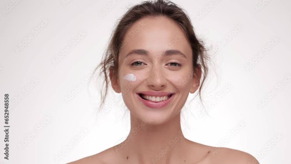 Female beauty procedures. Lady cream applying smear of pampering cream on her cheek and smiling to the camera, white studio background. Skin care concept. Cosmetic Concept. Female beauty procedures. Lady cream applying smear of pampering cream on her cheek and smiling to the camera, white studio background. Skin care concept. Cosmetic Concept.
