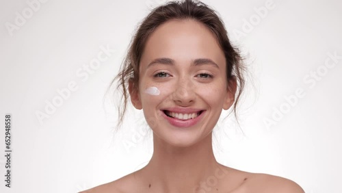 Female beauty procedures. Lady cream applying smear of pampering cream on her cheek and smiling to the camera, white studio background. Skin care concept. Cosmetic Concept.
