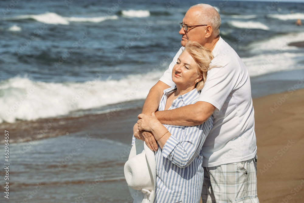 An elderly couple stand hugging on the beach by the sea, look at the waves, relax.