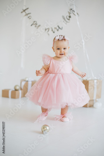 Playful little girl wearing finest party dress standing alone in studio adorned with festive decorations. Charming caucasian baby girl with sincere emotions celebrating birthday party.