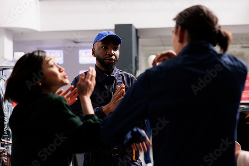 Calm polite African American male security officer managing crowd in shopping mall during sales, asking two angry mad shoppers to remain calm during Black Friday. People bargain hunters fighting