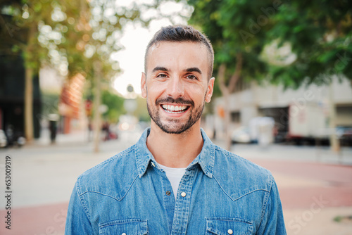 Close up portrait of handsome guy with perfect white teeth smiling and looking at camera standing outdoors. Front view of young man with friendly and positive expression. Happy adult male staring
