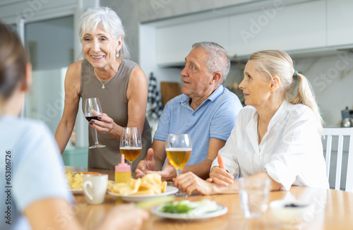Group of joyful elderly women and man gathered around cozy kitchen table, chatting amiably while drinking beer and wine with snacks. Concept of socialization of older adults