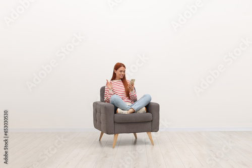 Happy young woman having video chat via smartphone while sitting in armchair near white wall indoors