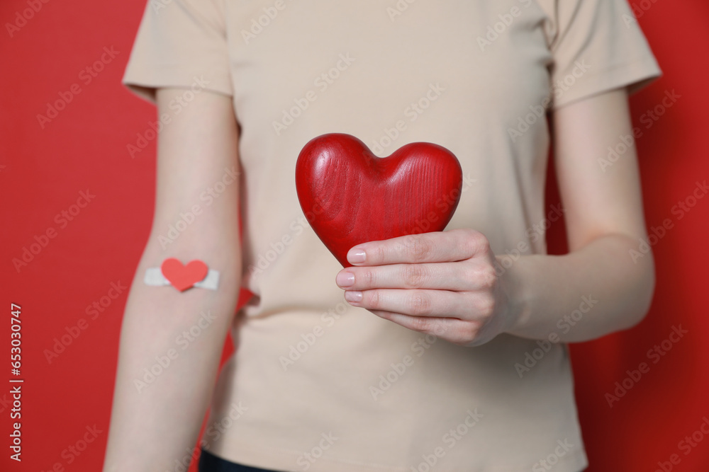 Blood donation concept. Woman with adhesive plaster on arm holding red heart against color background, closeup Blood donation concept. Woman with adhesive plaster on arm holding red heart against color background, closeup