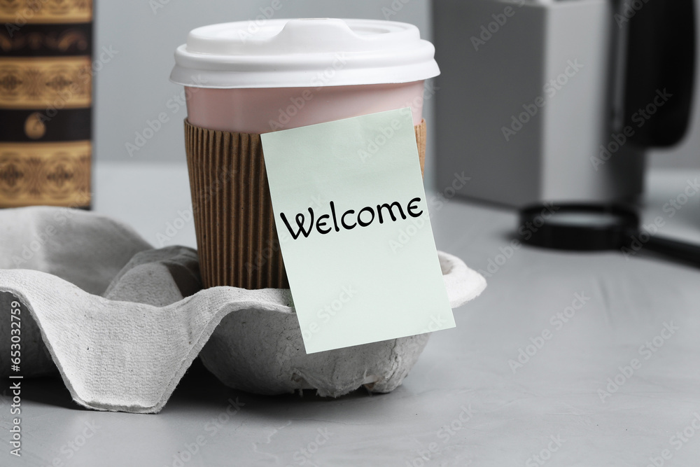 Sticky note with word Welcome attached to paper cup of coffee on office desk