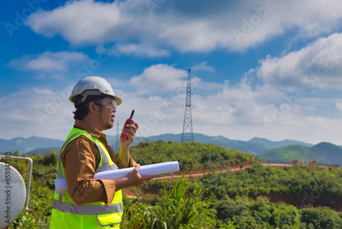 Engineer manager man with white helmet and hold construction paper plan and fm handheld transceiver or walkie talkie look back to camera and stand in front of mountain. photo