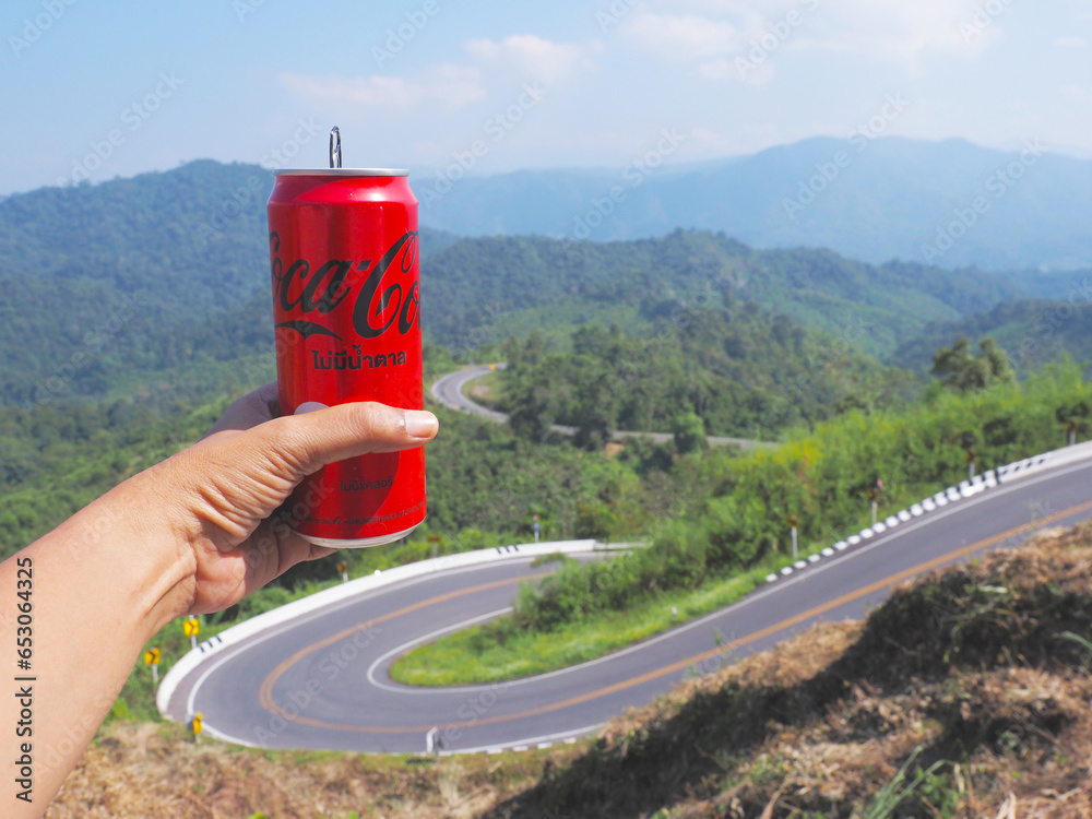 Fototapeta premium Hand holding red can of Coca-Cola soft drink at viewpoint over curve street on mountains background.