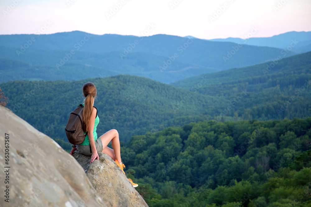 Tired hiker girl relaxing on rocky mountain top enjoying evening nature during travelling on wilderness trail. Lonely woman traveler traversing high hilltop route. Healthy lifestyle concept