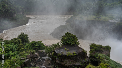 View from the observation deck of the Iguazu River. The riverbed bends between the steep banks. Green tropical vegetation on wet rocks. Spray and fog all around. Argentina. Iguazu Falls.