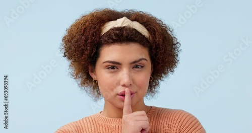 Whisper, face and woman with finger on lips in studio for secret, news or hush on blue background. Gossip, portrait and lady model with privacy, confidential or emoji for drama, announcement or mute photo