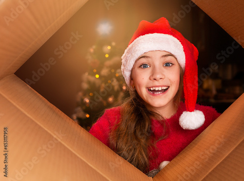 Little girl in Santa hat opening a cardboard box and looking at camera