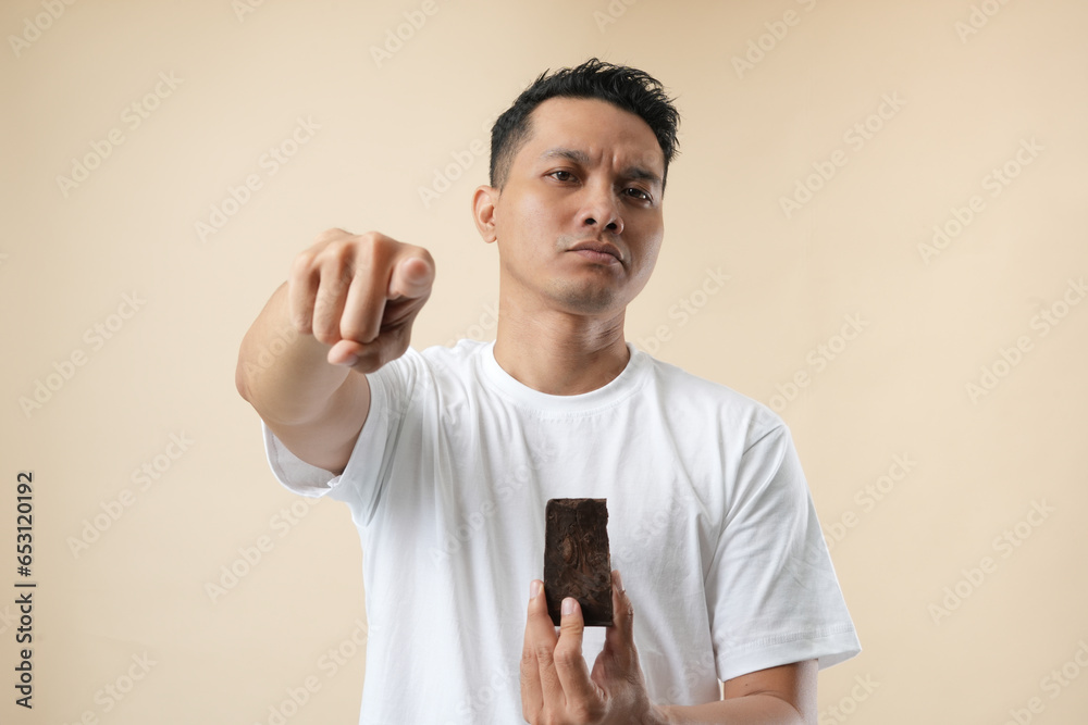 A young Asian man with acne problem holding a chocolate bar on a beige background. Skin allergy concept. A young Asian man with acne problem holding a chocolate bar on a beige background. Skin allergy concept.