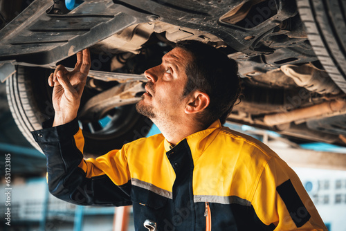 Vehicle mechanic conduct car inspection from beneath lifted vehicle. Automotive service technician in uniform carefully diagnosing and checking car's axles and undercarriage components. Oxus photo