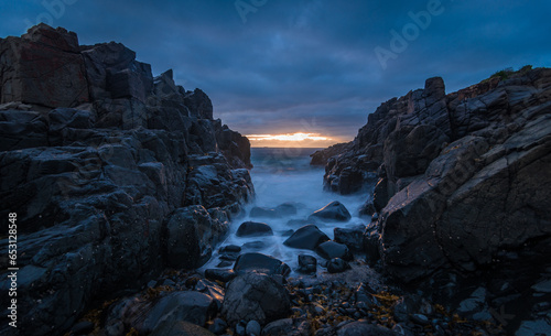A shot through a gap in the rocks of the old Bombo blue metal quarry looking towards the sun rising over the water running over the rocks. 