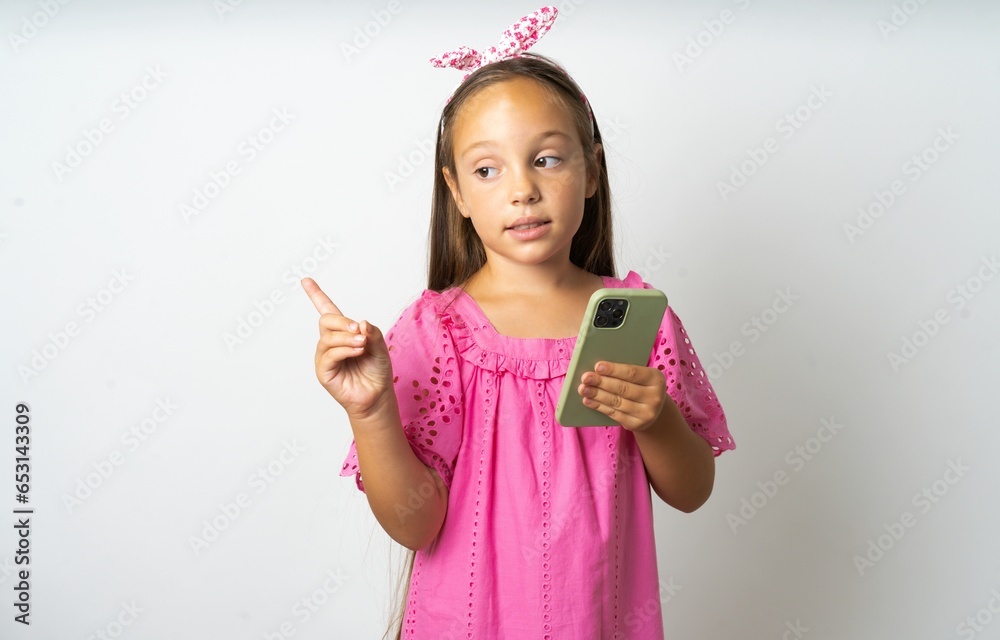 Smiling beautiful kid girl wearing pink dress holding mobile phone and pointing aside at copyspace. Smiling beautiful kid girl wearing pink dress holding mobile phone and pointing aside at copyspace.