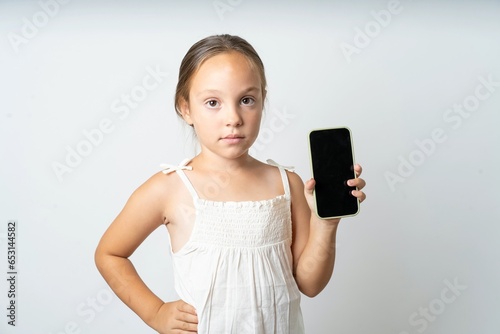beautiful kid girl wearing white dress holds new mobile phone and looks mysterious aside shows blank display of modern cellular
