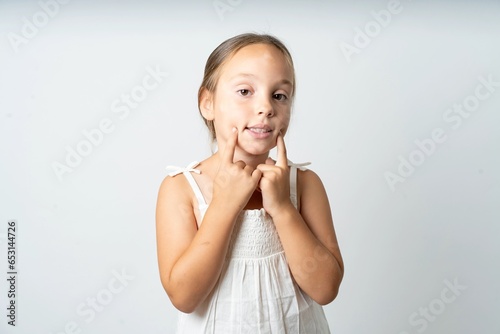Happy beautiful kid girl wearing white dress with toothy smile, keeps index fingers near mouth, fingers pointing and forcing cheerful smile