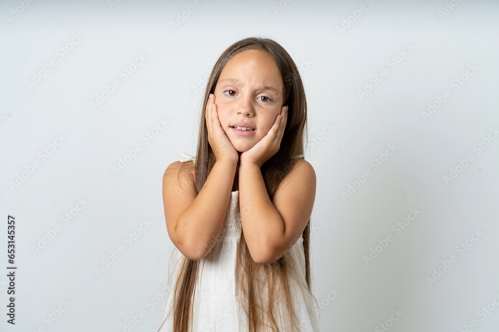 Upset beautiful kid girl wearing white dress touching face with two hands Upset beautiful kid girl wearing white dress touching face with two hands