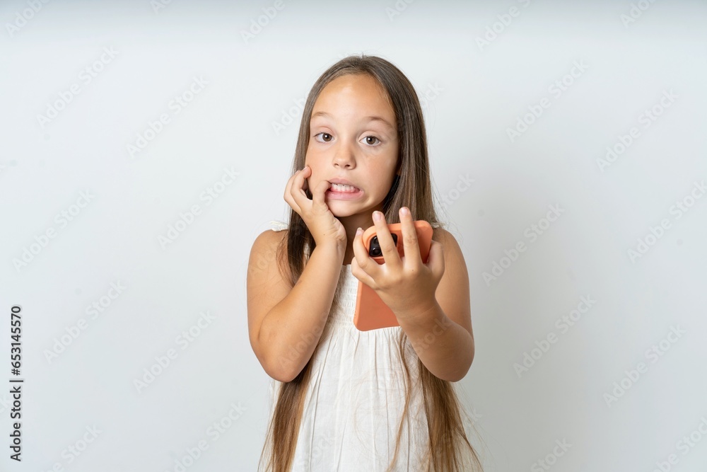 Afraid funny beautiful kid girl wearing white dress holding telephone and bitting nails Afraid funny beautiful kid girl wearing white dress holding telephone and bitting nails