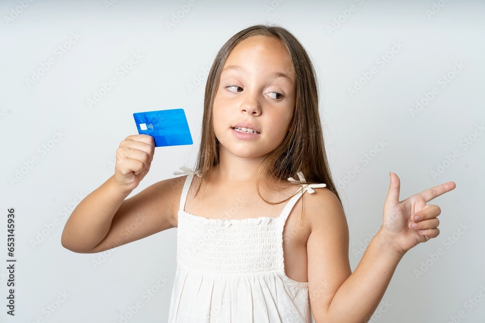 Curious smiling beautiful kid girl wearing white dress showing plastic bank showing finger copyspace Curious smiling beautiful kid girl wearing white dress showing plastic bank showing finger copyspace