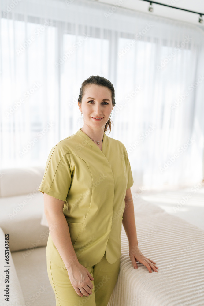 Medium shot vertical portrait of smiling female masseuse therapist in uniform standing posing by working couch looking at camera with friendly expression, on background of window in wellness center. Medium shot vertical portrait of smiling female masseuse therapist in uniform standing posing by working couch looking at camera with friendly expression, on background of window in wellness center.