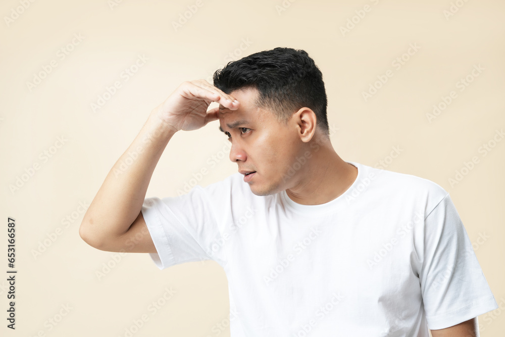A young Asian man wearing a white t-shirt over an isolated cream background, happy and looking far away with a hand over his head. Searching concept. A young Asian man wearing a white t-shirt over an isolated cream background, happy and looking far away with a hand over his head. Searching concept.
