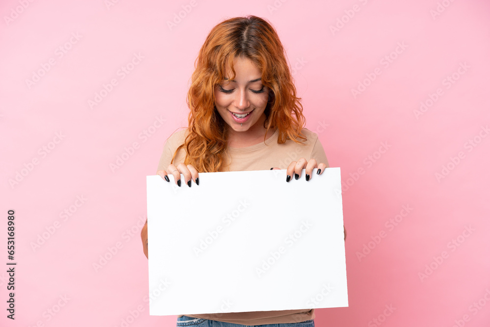 Naklejka premium Young caucasian woman isolated on pink background holding an empty placard and looking it