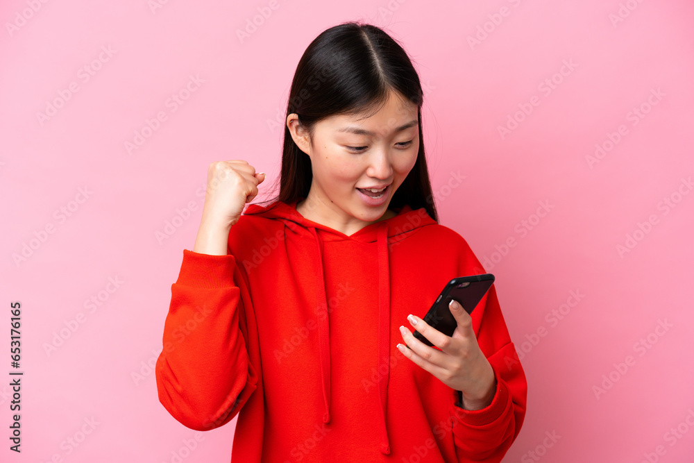 Young Chinese woman isolated on pink background using mobile phone and doing victory gesture