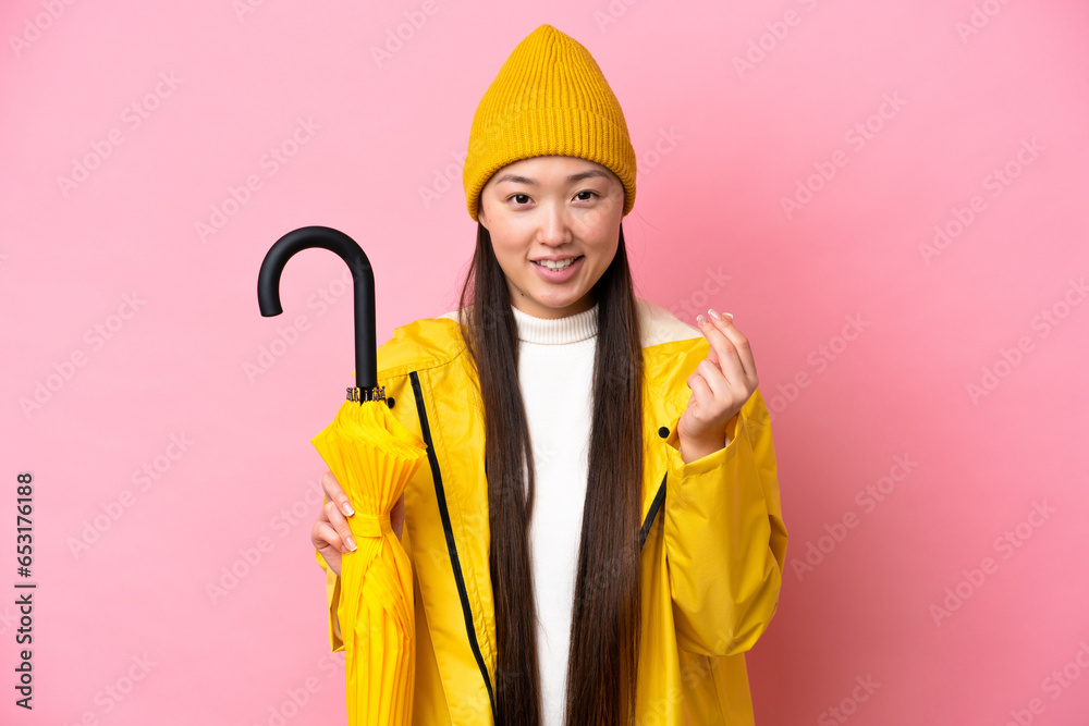 Young Chinese woman with rainproof coat and umbrella isolated on pink background making money gesture Young Chinese woman with rainproof coat and umbrella isolated on pink background making money gesture