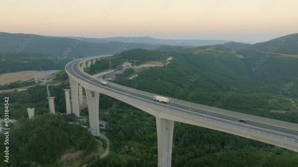 Trucks driving on road bridge at sunset with mountains skyline aerial tracking shot. Shipment lorry riding on highway and transporting cargo drone view Trucks driving on road bridge at sunset with mountains skyline aerial tracking shot. Shipment lorry riding on highway and transporting cargo drone view