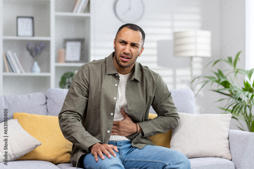 A young African American man is suffering from abdominal pain, stomach cramps and constipation. He is sitting on the sofa and holding his stomach, grimacing in pain A young African American man is suffering from abdominal pain, stomach cramps and constipation. He is sitting on the sofa and holding his stomach, grimacing in pain