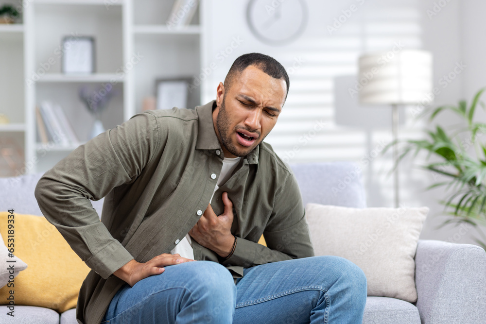 A young African-American man is suffering from a heart attack, sitting on the sofa at home and grimacing, holding his hand to his chest