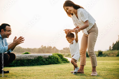 A happy mother and father helping their toddler take her first steps in the green grass of the park. baby learning concept