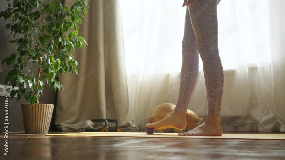 Woman warms up her feet with a massage ball on a sports mat at home in the room, close-up of her legs. Woman warms up her feet with a massage ball on a sports mat at home in the room, close-up of her legs.