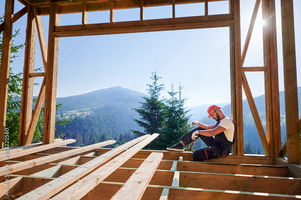 Carpenter constructing wooden frame two-story house near the forest. Bearded man holding hammer, dressed in protective helmet and overalls. Concept of ecological modern construction. Carpenter constructing wooden frame two-story house near the forest. Bearded man holding hammer, dressed in protective helmet and overalls. Concept of ecological modern construction.