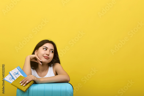 Cute dreamy brunette girl affectionately looks away, holds plane tickets in her hands. An Asian kopeck piece tourist in anticipation of the departure of the plane folded her hands on a blue suitcase