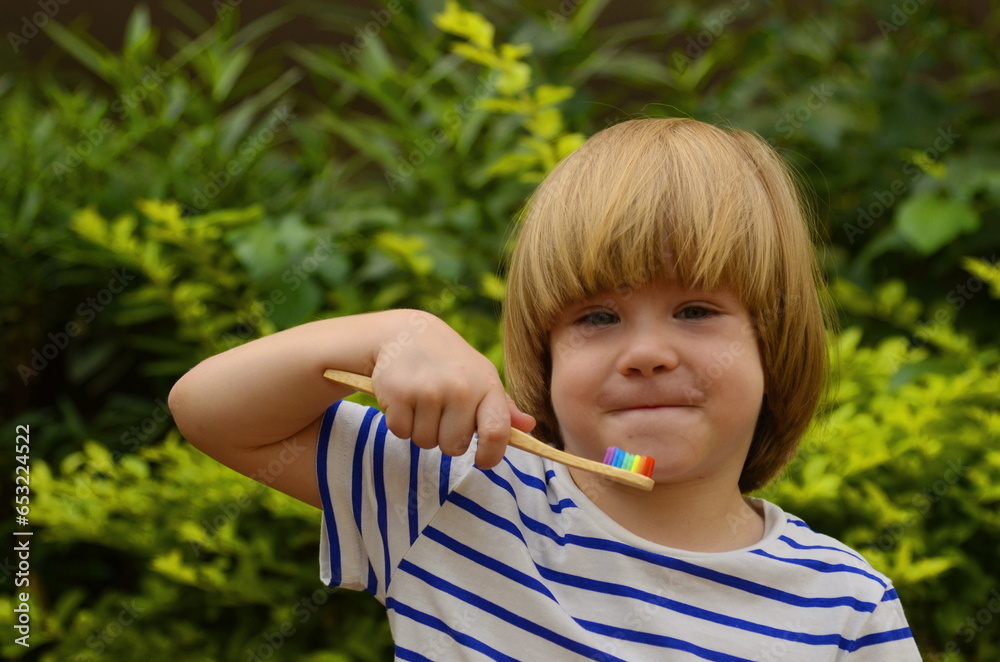 A little boy brushes his teeth in the garden in nature, on the street. Oral hygiene. Ecological wooden brush. Eco-friendly. Multi-colored toothbrush, green foliage in the background. A little boy brushes his teeth in the garden in nature, on the street. Oral hygiene. Ecological wooden brush. Eco-friendly. Multi-colored toothbrush, green foliage in the background.