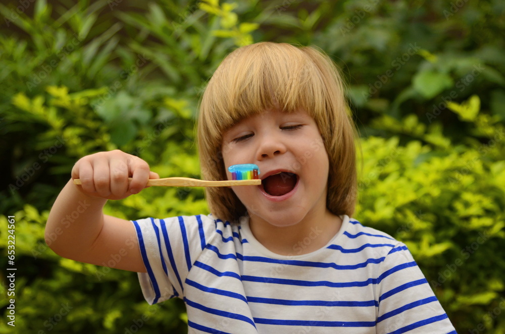 A little boy brushes his teeth in the garden in nature, on the street. Oral hygiene. Ecological wooden brush. Eco-friendly. Multi-colored toothbrush, green foliage in the background. A little boy brushes his teeth in the garden in nature, on the street. Oral hygiene. Ecological wooden brush. Eco-friendly. Multi-colored toothbrush, green foliage in the background.