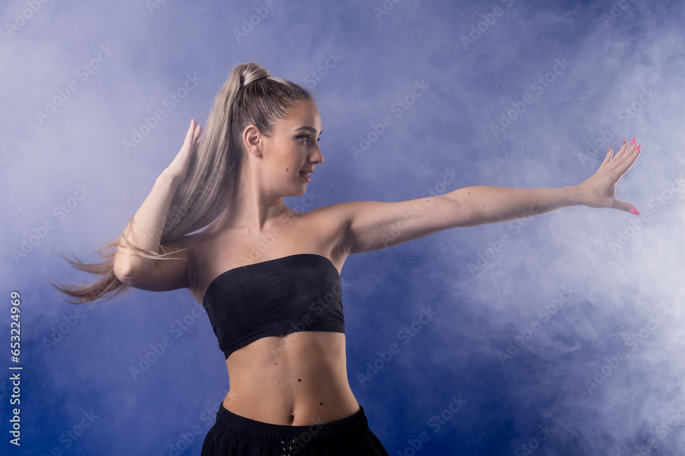 Fototapeta premium Beautiful blond girl in black outfit dancing zumba against smoke fog and blue background.