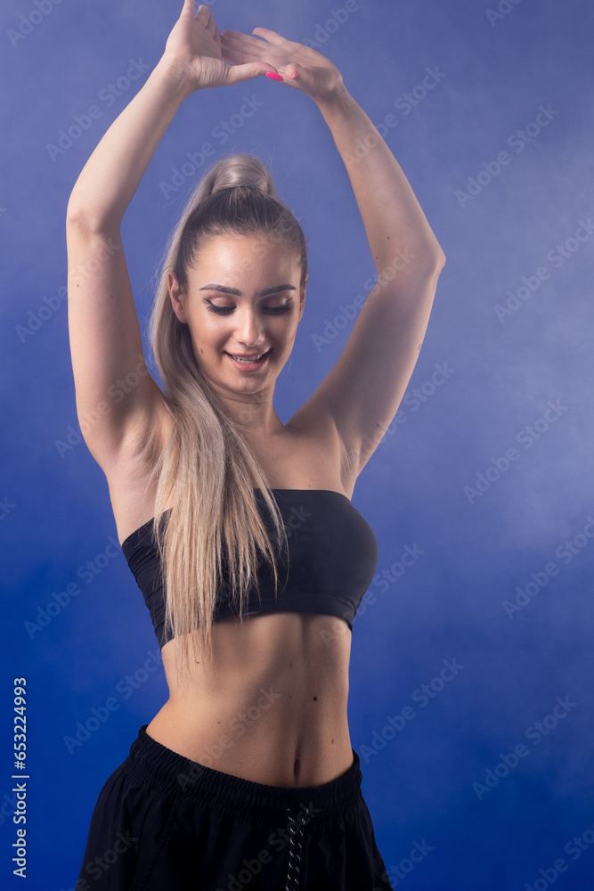 Beautiful blond girl in black outfit dancing zumba against smoke fog and blue background.