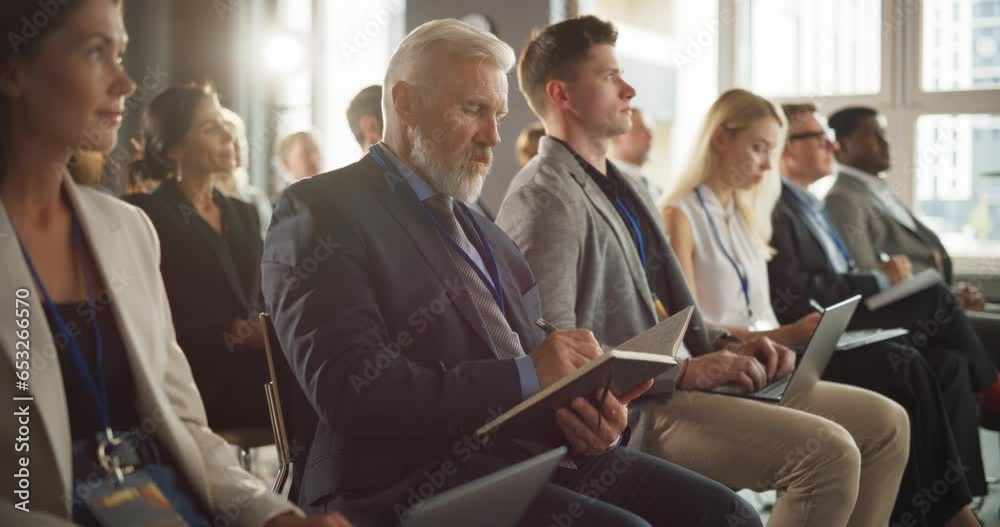 Senior Caucasian Man Sitting in a Crowded Audience at a Business Forum. Corporate Delegate Taking Notes. Successful Male CEO Attending Business Conference, Listening To Presentation. Senior Caucasian Man Sitting in a Crowded Audience at a Business Forum. Corporate Delegate Taking Notes. Successful Male CEO Attending Business Conference, Listening To Presentation.