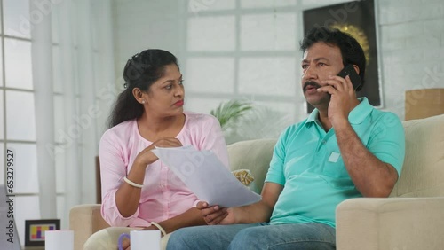 Serious Indian middle aged man with wife talking on mobile phone by holding property documents at home - concept of conversation, investment planning and togetherness