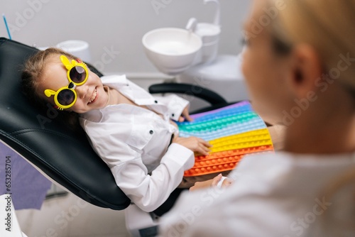 Side view of cheerful little child girl in funny eyeglasses playing with colorful pop it toy, sitting in dental chair during examining teeth from pediatric dentist. Concept of children teeth treatment