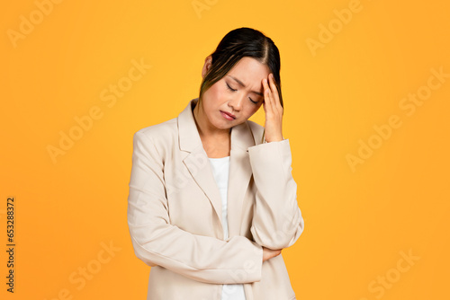 Sad young asian woman in suit presses hand to head, suffering from headache