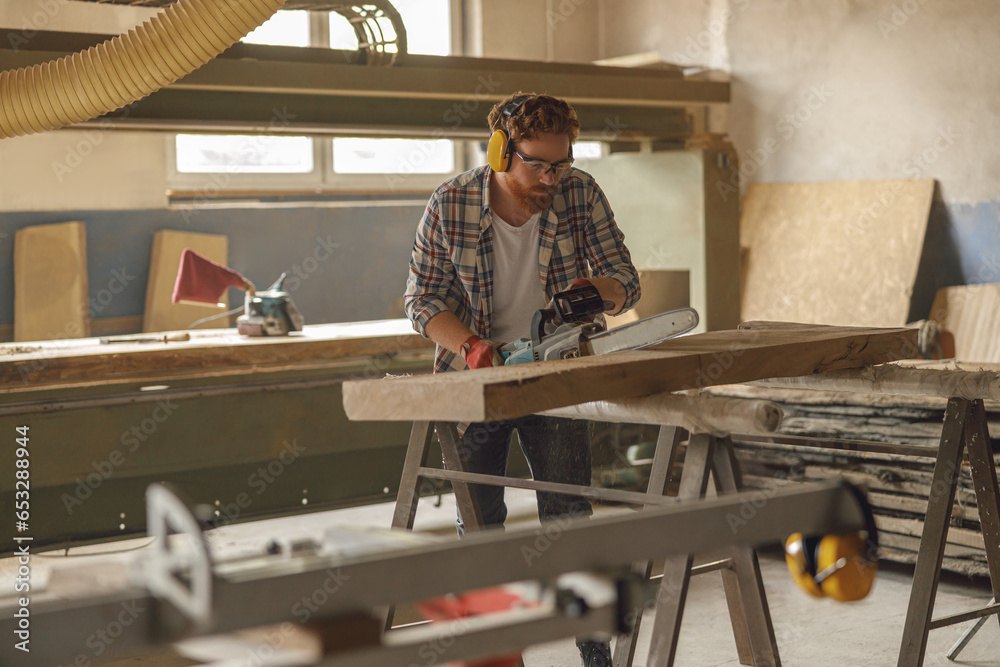 Fototapeta premium Professional carpenter in glasses is cutting wood with chainsaw on carpentry workshop
