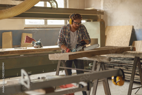 Professional carpenter in glasses is cutting wood with chainsaw on carpentry workshop