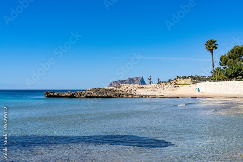 Scenic view of the calm, clear shoreline at Platja de l'Ampolla beach in Moraira, Spain, looking across the bay toward a rocky point and Calpe's Peñón de Ifach under a clear blue sky.